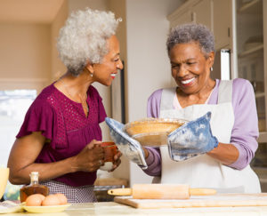 A smiling Black woman with gray hair holds a pie she baked while another Black woman smiles back at her.