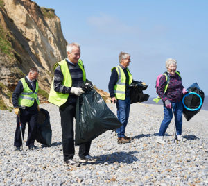 A group of four older white men and women wearing reflective vests and carrying trash bags to pick up litter on the beach.
