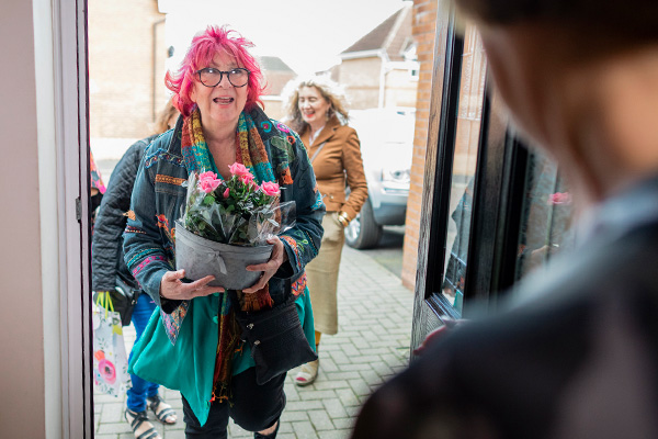 An older woman with colorful hair bringing flowers through a friend’s front door while smiling